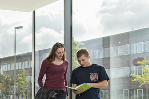 Vier Personen stehen in einem Uni-Gebäude am Fenster. Zwei von ihnen unterhalten sich, die beiden anderen schauen gemeinsam in ein Buch.