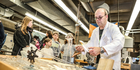 Ein Mann steht in einem weißen Laborkittel an einem Tisch im Labor und Kinder stehen um den Tisch herum.