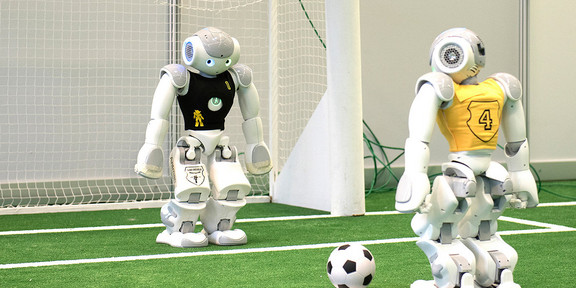 Soccer robot penalty kick A robot with a black jersey stands in a soccer goal and a second robot stands with a ball at the penalty spot.