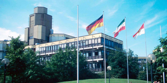 Altes Physikgebäude: Ein braun-gelbes Gebäude mit drei Stockwerken und einem hohen Turm links hinten. Davor eine Wiese und ein Platz mit Bänken und vier Fahnenmasten, an denen eine Deutschland-Fahne, eine NRW-Fahne und eine Dortmund-Fahne.