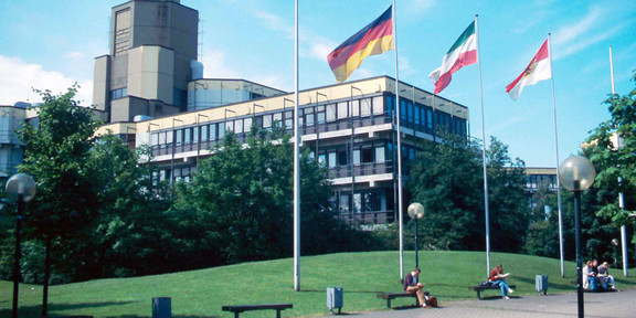 Altes Physikgebäude: Ein braun-gelbes Gebäude mit drei Stockwerken und einem hohen Turm links hinten. Davor eine Wiese und ein Platz mit Bänken und vier Fahnenmasten, an denen eine Deutschland-Fahne, eine NRW-Fahne und eine Dortmund-Fahne.