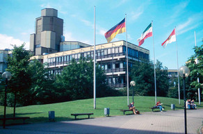 Altes Physikgebäude: Ein braun-gelbes Gebäude mit drei Stockwerken und einem hohen Turm links hinten. Davor eine Wiese und ein Platz mit Bänken und vier Fahnenmasten, an denen eine Deutschland-Fahne, eine NRW-Fahne und eine Dortmund-Fahne.